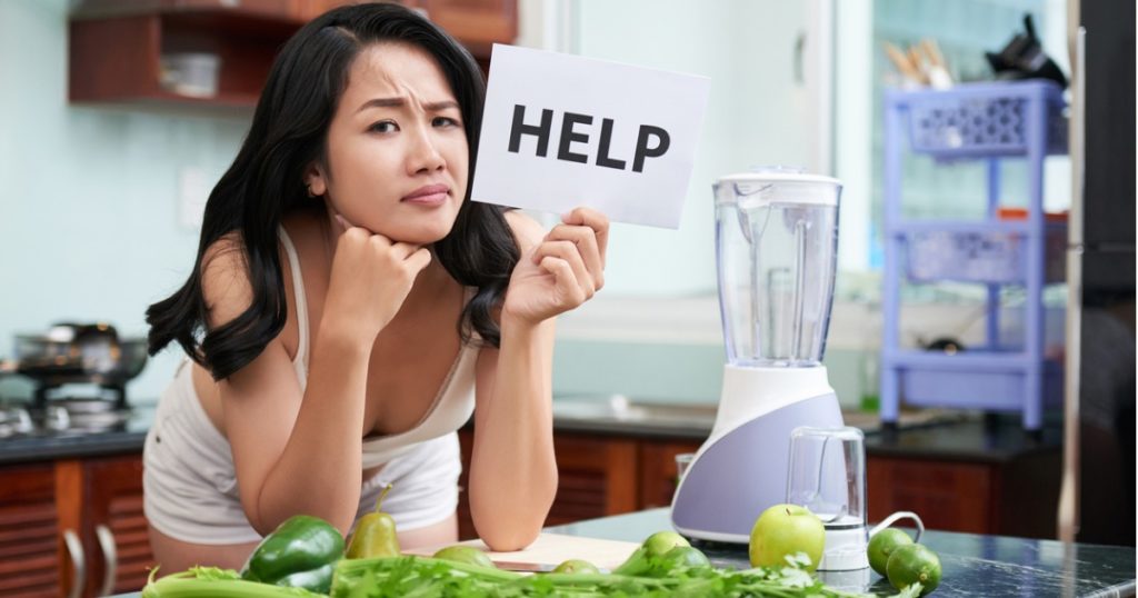 一名妇女靠在厨房柜台上，桌上摆满了水果和蔬菜，手里举着一个白色的牌子，上面写着“救命”。