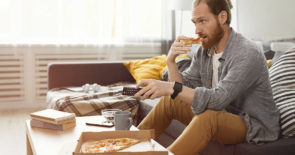一名男子坐在沙发上看电视，一边吃着纸板箱里的披萨，一边看起来很焦虑。